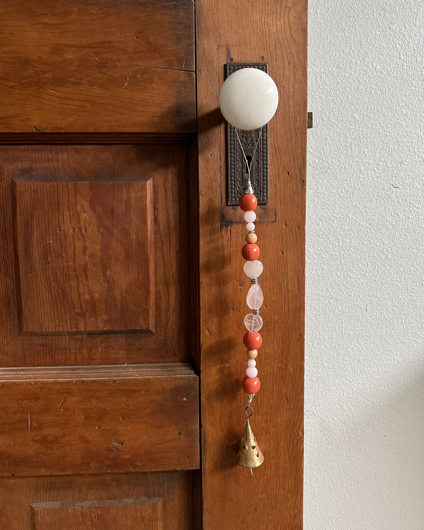 Single strand of wooden beads, rose quartz, and a golden bell hang from an antique doorknob.