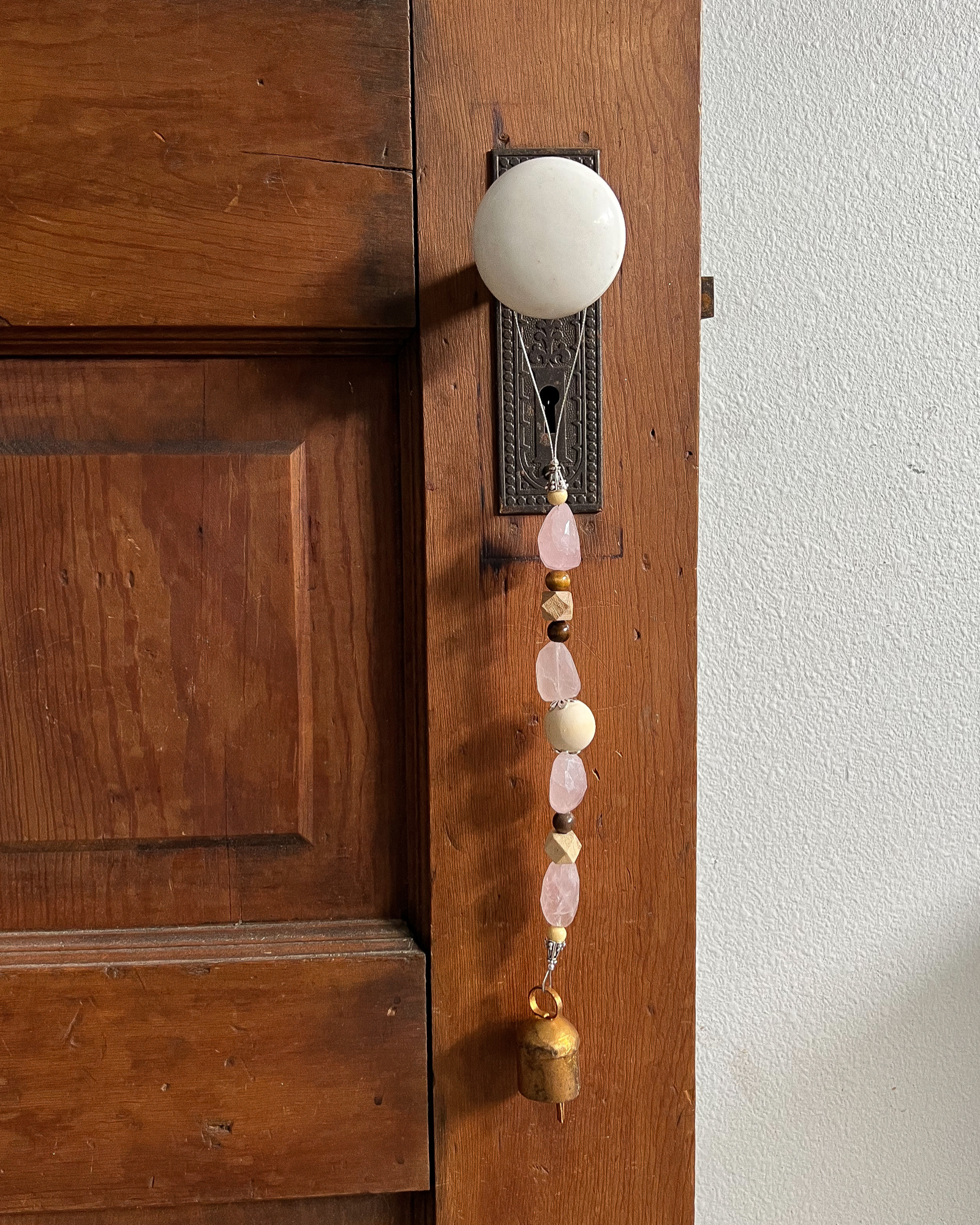 Single strand of wooden beads, rose quartz, and a golden bell hang from an antique doorknob.