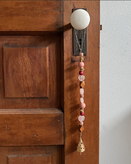Single strand of wooden beads, rose quartz, and a golden bell hang from an antique doorknob.