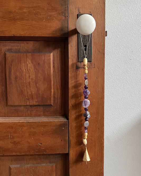 Single strand of wooden beads, metal charms, amethyst crystals, and a golden bell hanging from an antique doorknob.