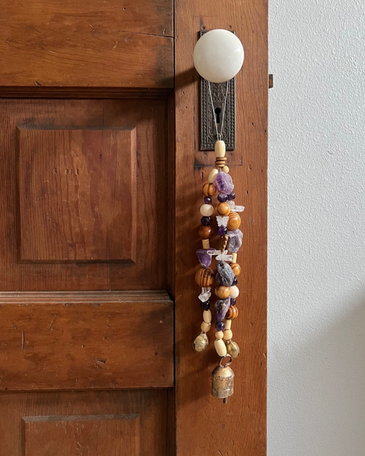 Triple strand of wooden beads, amethyst crystals, a single clear butterfly bead, and three golden bells hanging on an antique doorknob.