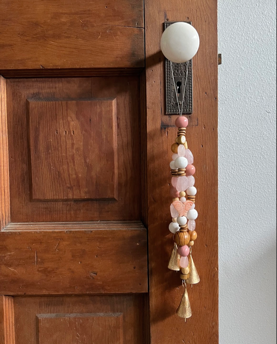 Triple strands of wooden beads, rose quartz crystals, a single rose quartz butterfly, and three golden bells hanging from an antique doorknob.